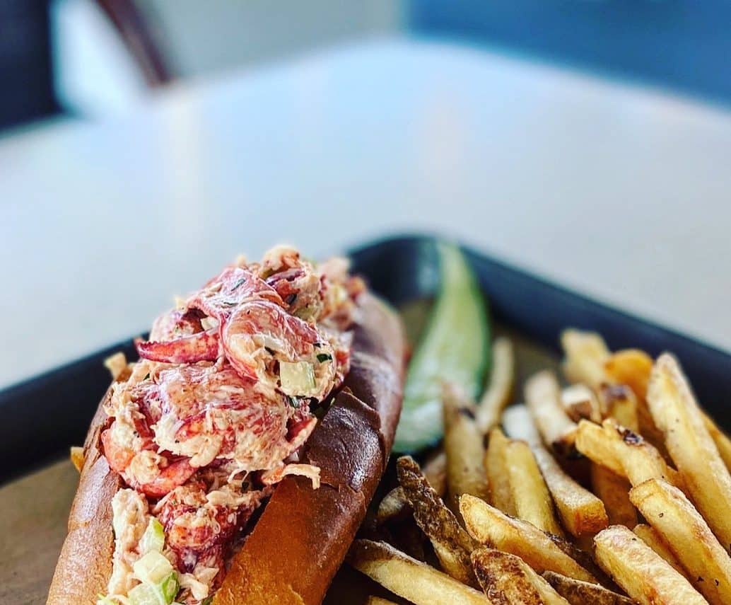 A plate with a large lobster salad roll, french fries and a pickle.