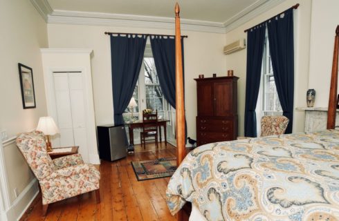 Spacious guest-room with original wooden floors and a four post bed with a blue and tan cover and a wooden armoire.
