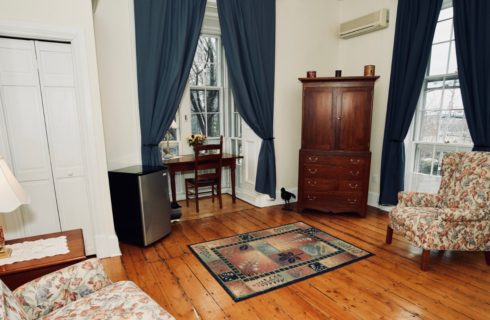 Room with large window with blue drapes, a wooden armoire, mini-fridge and desk.