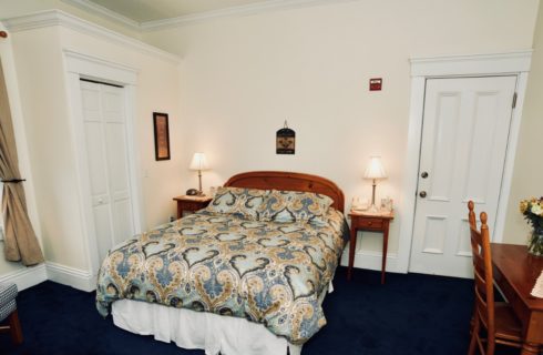 Guest-room with a queen bed featuring a blue and tan comforter, two nightstands with lamps, and a wooden desk