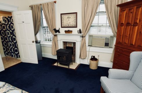 Guest-room with a wood stove, blue patterned wing chair, wooden armoire, and blue wall-to-wall carpeting.