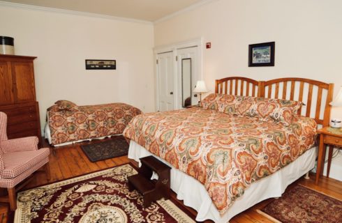 Bedroom with a king and twin bed with nigh stands and lamps, red wingback chair, and a wooden armoire.
