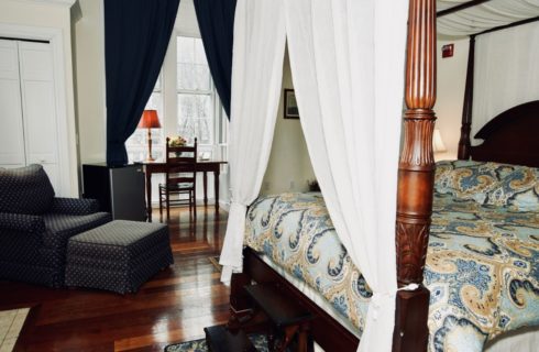 Wooden canopy bed in a bedroom with a blue chair and ottoman and a desk under a large window.