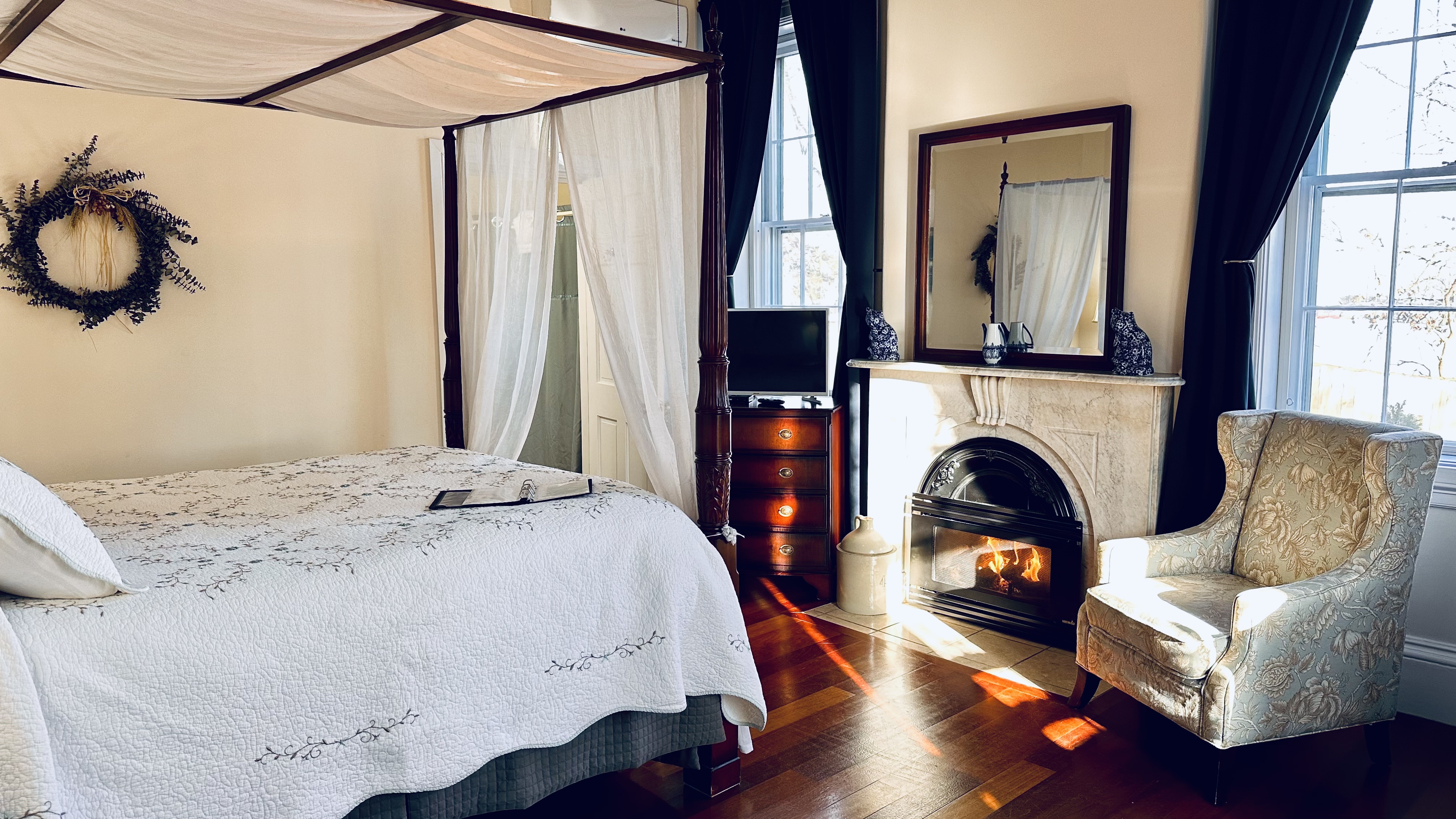 A queen canopy bed with a white bed spread facing a marble fireplace and an upholstered chair/