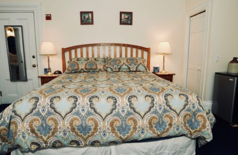 A king bed made up in a blue covering, 2 nightstands with lamps, and a small refrigerator.