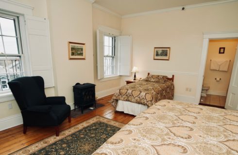 King and twin bed in a room facing shuttered windows, green wingback chair, and a wood stove