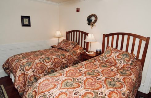 A cozy room featuring two twin beds made up in red patterned comforters featuring two end tables with lamps.