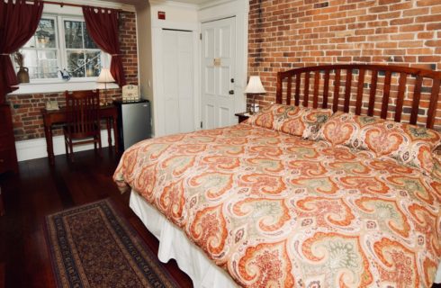 Big king sized bed in a room with a brick accent wall, wooden desk, and small refrigerator.