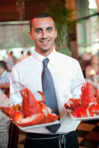 A gentleman waiter holding to plates of large cooked red lobsters.