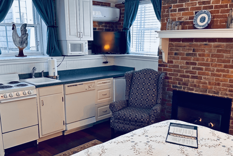 A room with a bed, kitchenette, brick fireplace, and a chair.