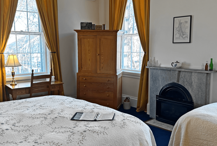 A room with 2 beds, a mini fridge, desk with a chair, armoire, and marble fireplace.