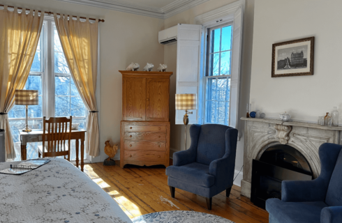 A room with a bed, mini fridge, desk with chair, armoire, 2 chairs, and a marble fireplace.