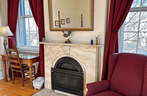 A room with a chair, marble fireplace, and desk with chair.