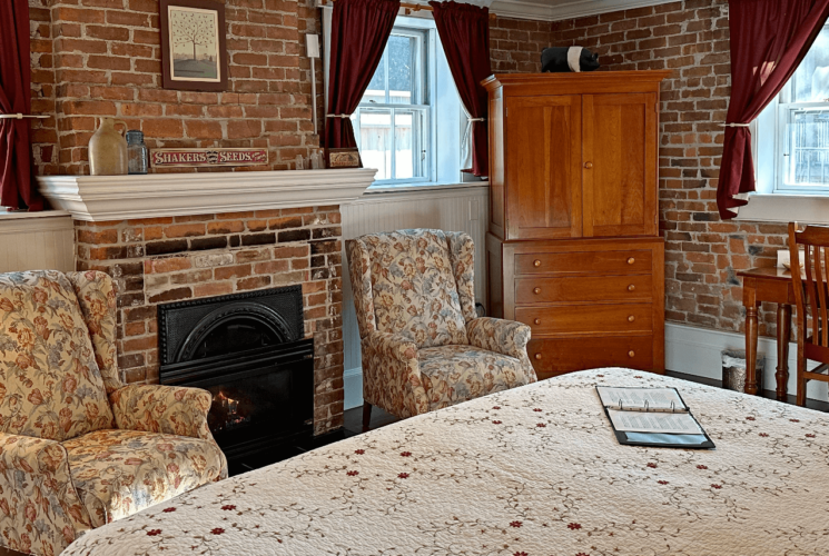 A room with a bed, brick fireplace, 2 chairs, armoire, and desk with a chair.