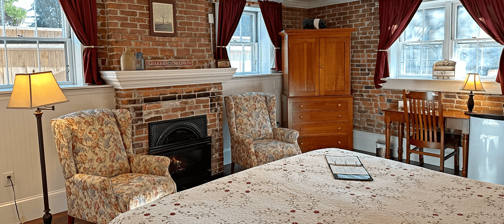 A room with a bed, brick fireplace, 2 chairs, armoire, and desk with a chair.