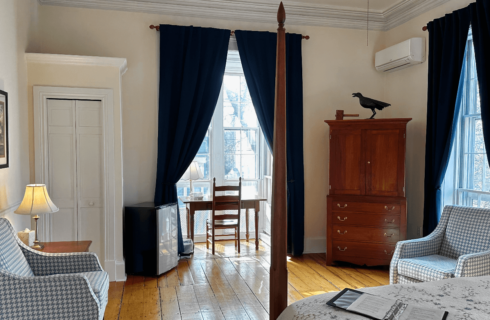 A room with a 4 poster bed, desk with chair, armoire, mini fridge, and a chair.