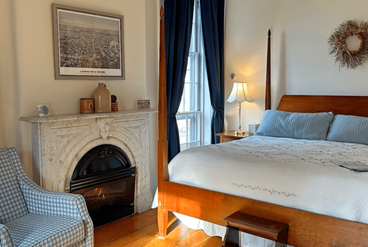 A room with a 4 poster bed, marble fireplace, and a chair.