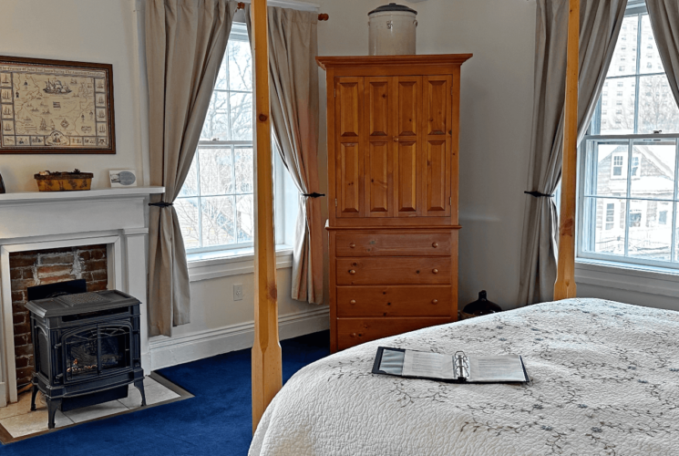 A room with a four-poster bed, a chair, wood stove, and an armoire.