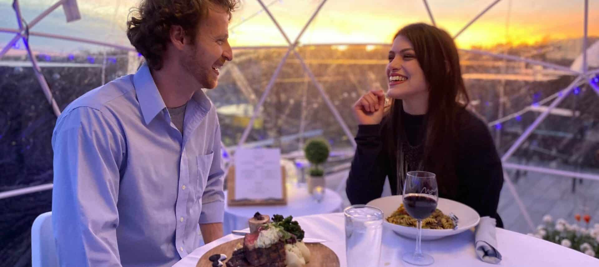 A smiling couple enjoying a meal inside an igloo while the sun sets behind them.