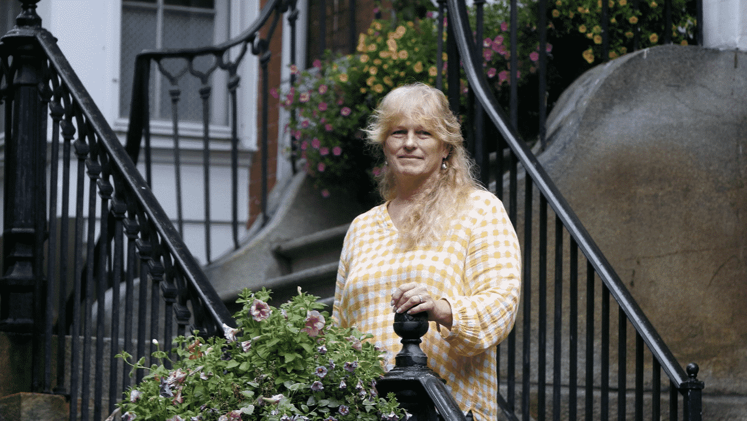 A woman wearing a yellow plaid top standing next to a beautiful flower box attached to a black iron railing.