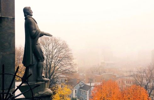 A statue a a colonial man looks over a town covered in fall foliage on a misty morning.