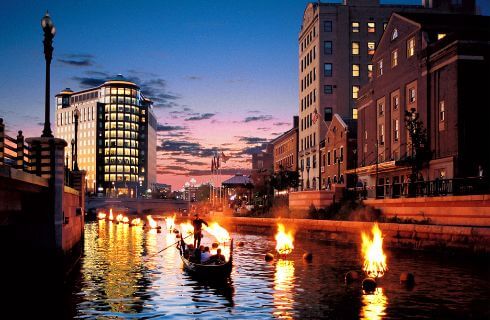 A gondola sails down the river next to a cityscape at dusk.