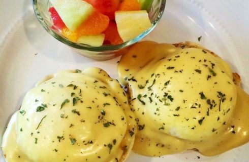 Eggs Benedict on a white plate with a bowl of fresh fruit salad.