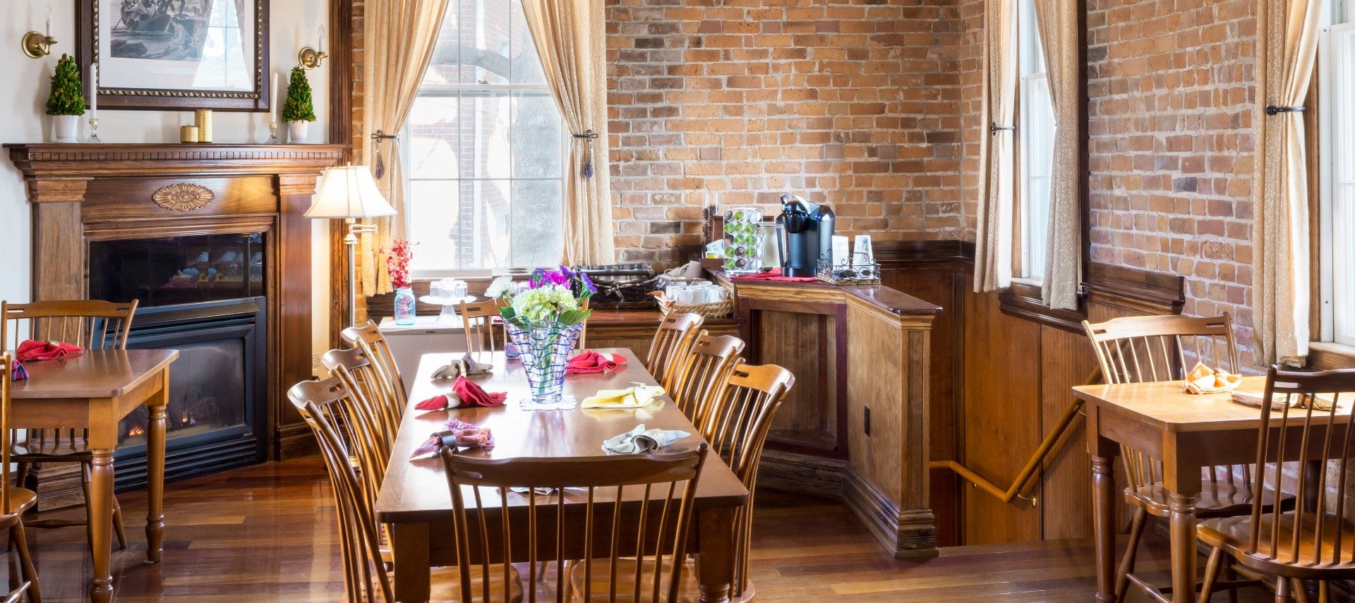 A large dining room with open seating facing a beautifully framed fireplace and large bright windows.