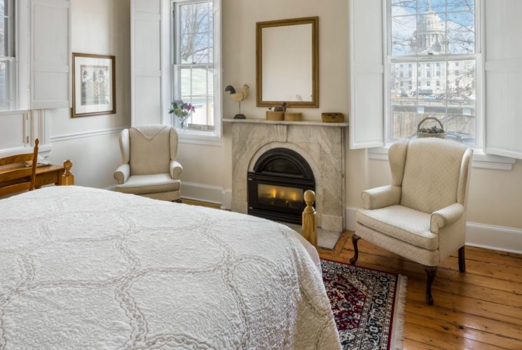 Oak four-post bed faces a fireplace and wing back chair under a large window next to an armoire.