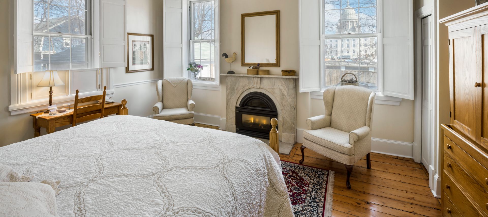 Oak four-post bed faces a fireplace and wing back chair under a large window next to an armoire.