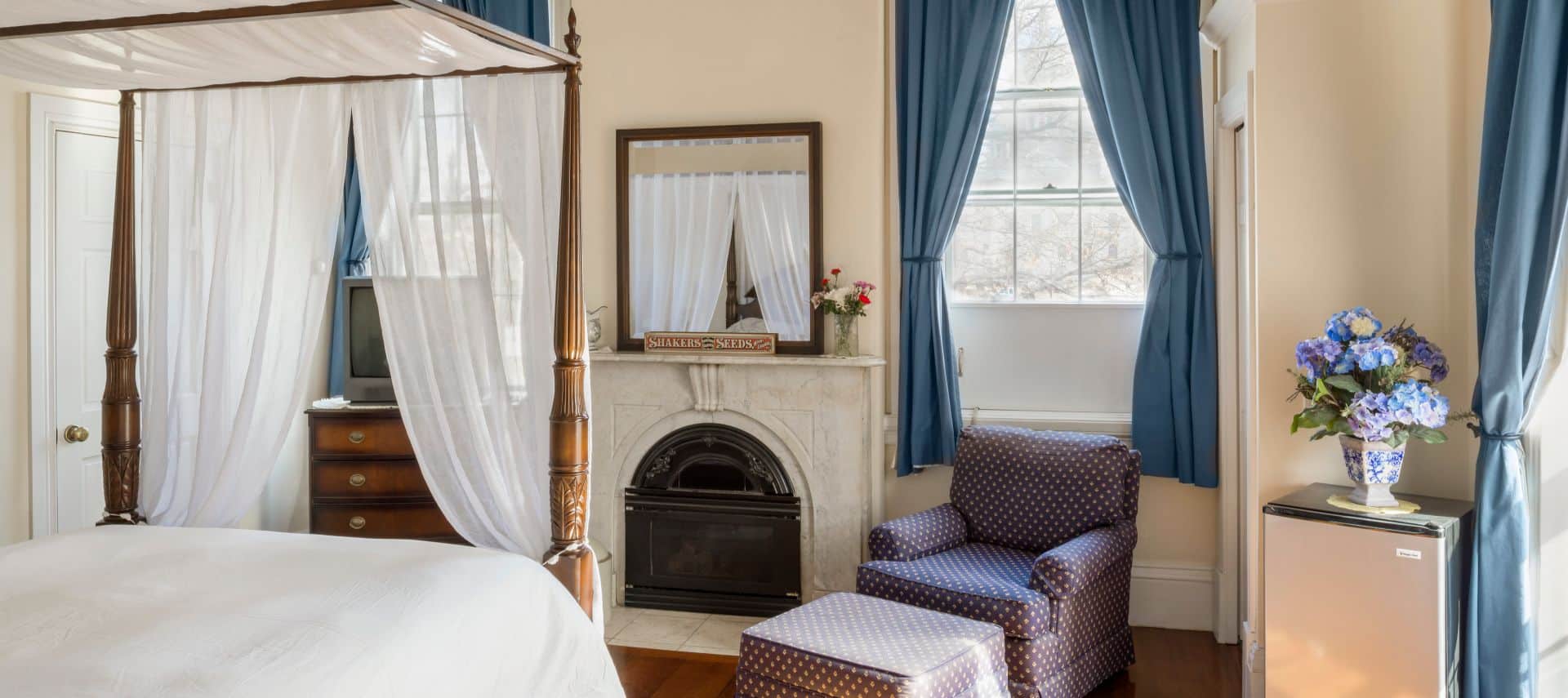 Queen canopy bed faces a marble fireplace with a mirror over it and a dresser with a TV.