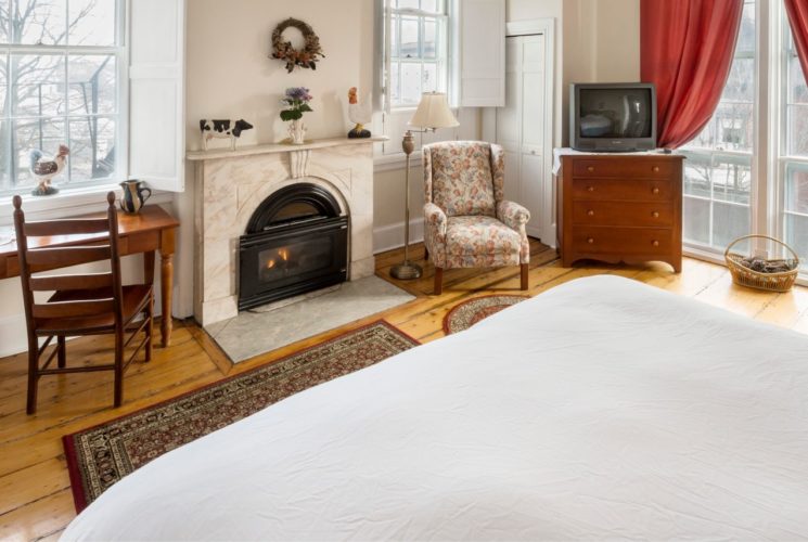 A King bed in a bedroom facing a wooden desk, fireplace, overstuffed chair, and large windows.