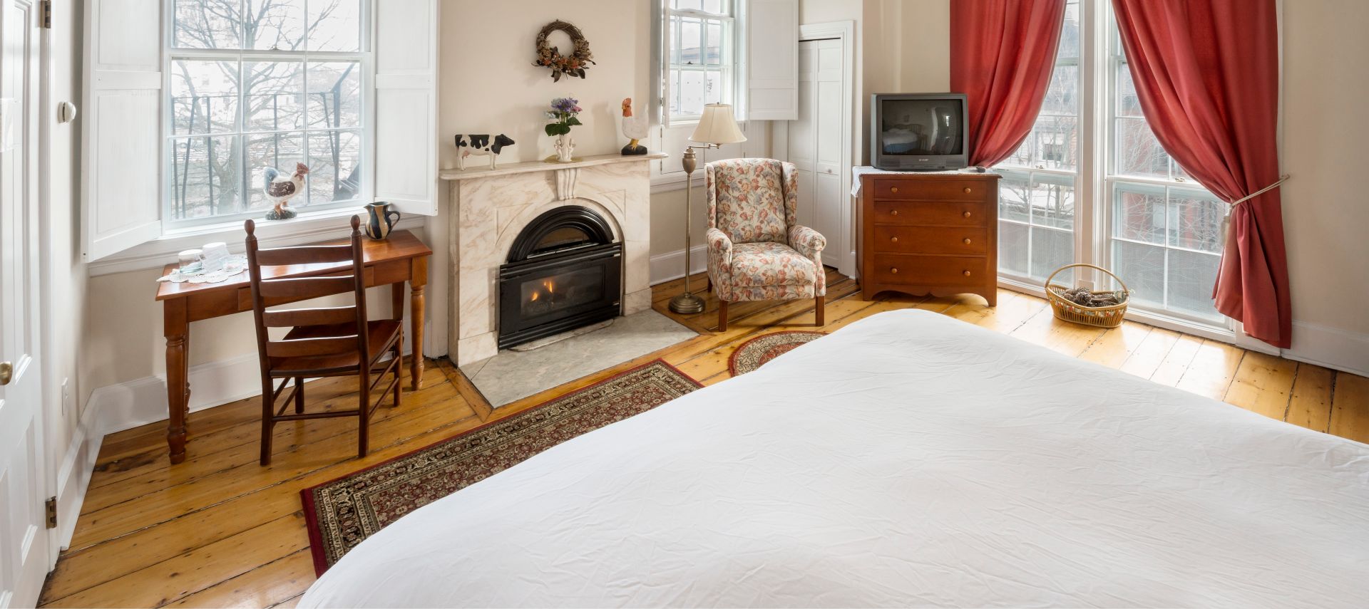 A King bed in a bedroom facing a wooden desk, fireplace, overstuffed chair, and large windows.