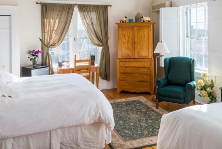 King and twin bed in a room with a wooden desk and armoire, wood stove, and tall windows.