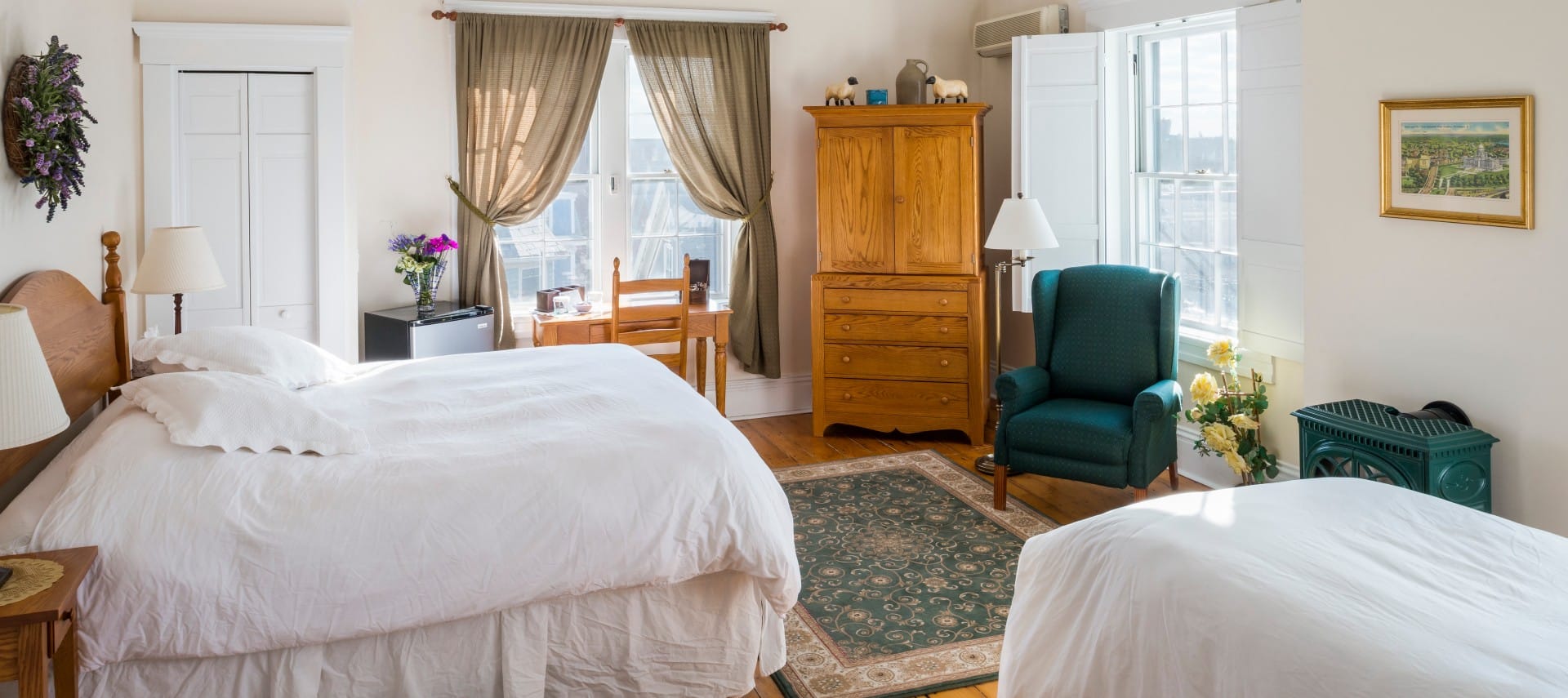 King and twin bed in a room with a wooden desk and armoire, wood stove, and tall windows.