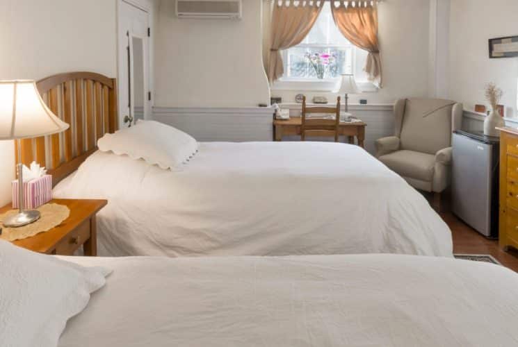 A cozy room featuring two twin beds made up in white comforters, wooden dresser with a tv, small refrigerator, beige wingback chair, and a wooden desk.