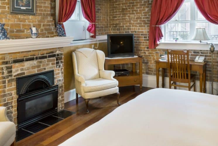 Bedroom with a brick fireplace, king sized bed, wooden tv stand and desk, two cream wingback chairs, and 3 bright windows.