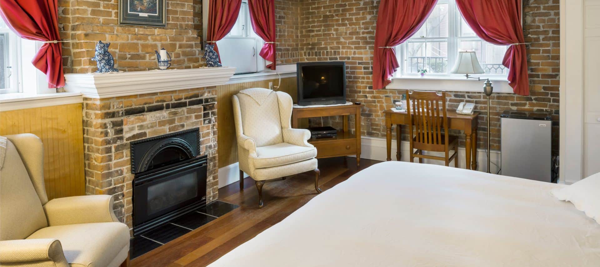 Bedroom with a brick fireplace, king sized bed, wooden tv stand and desk, two cream wingback chairs, and 3 bright windows.