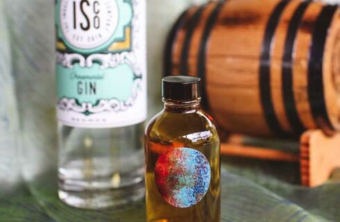 A tall colorful bottle of gin pictured infront of an old gin barrel.