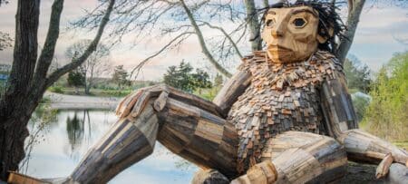 Large wooden troll sculpture by Thomas Dambo, made from recycled wood, sitting by a pond under trees