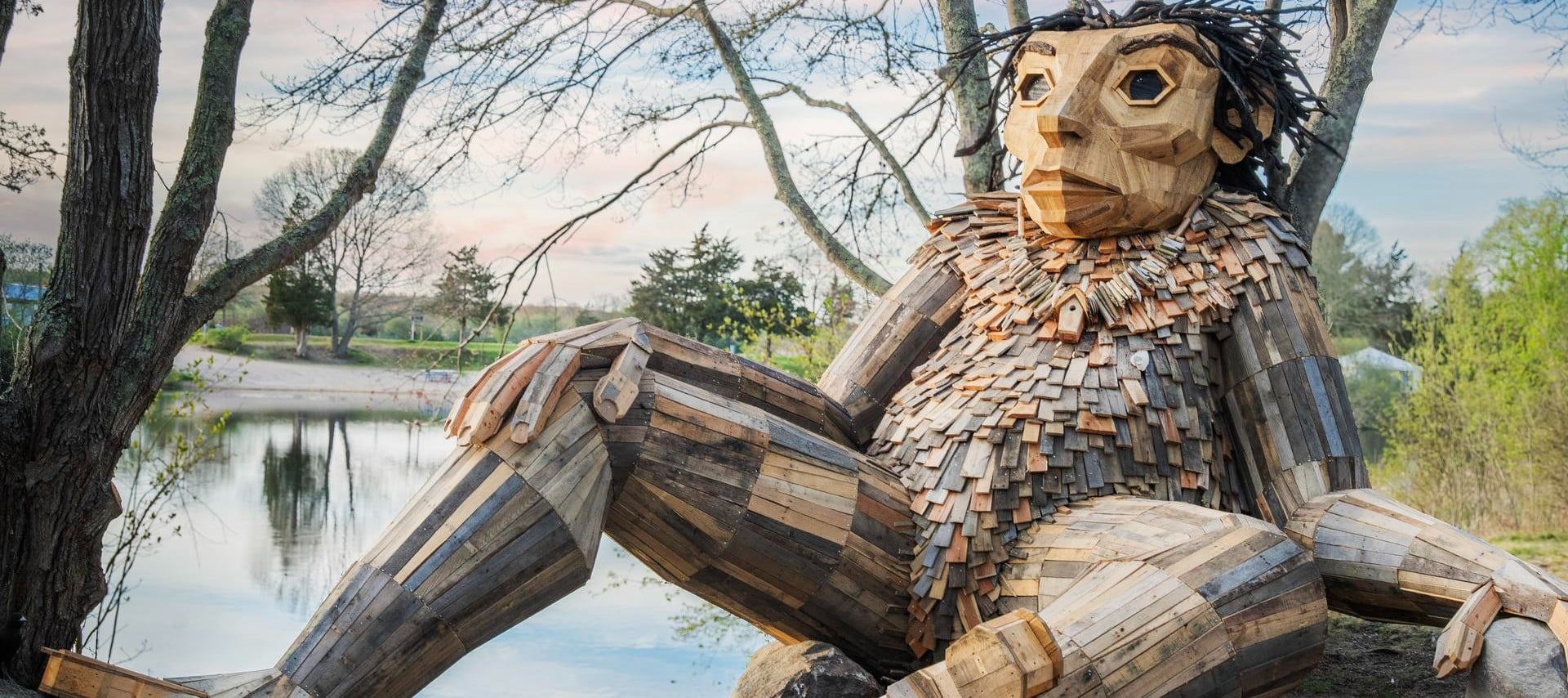 Large wooden troll sculpture by Thomas Dambo, made from recycled wood, sitting by a pond under trees